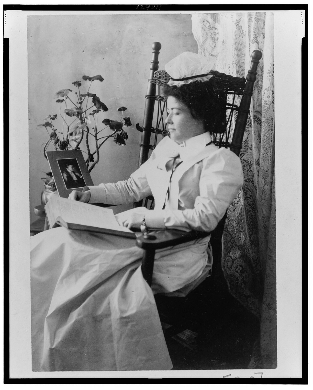 A nursing student in a starched white uniform and cap, seated in a rocking chair reading a book, photographed around 1900