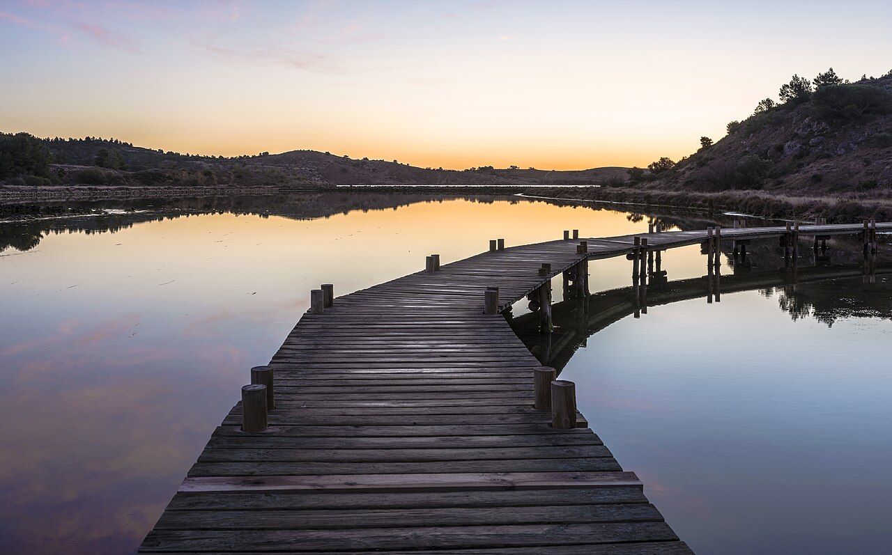 A weathered wooden boardwalk running out across a mirror-still lagoon at sunset, the far hills and sky doubled in the water