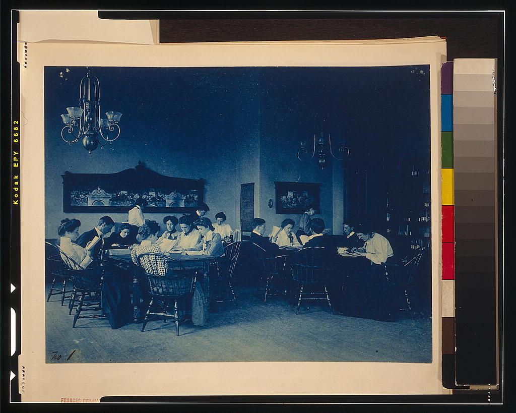 Students seated around a seminar table exchanging printed papers, one pointing at a passage while another writes in the margin
