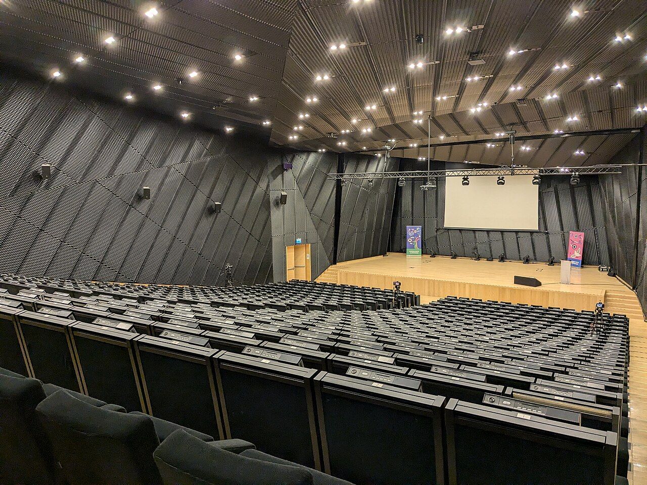 An empty auditorium seen from the stage, rows of seats facing a lectern and a projection screen